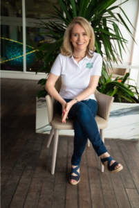 Claudia, the founder of Mighty Green who has blonde, mid-length hair, sitting on a beige coloured chair in front of some palm-type trees.