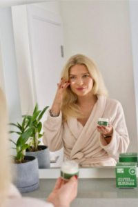 Blonde haired woman applying Mighty Green CBD Balm to her face in front of a large mirror.